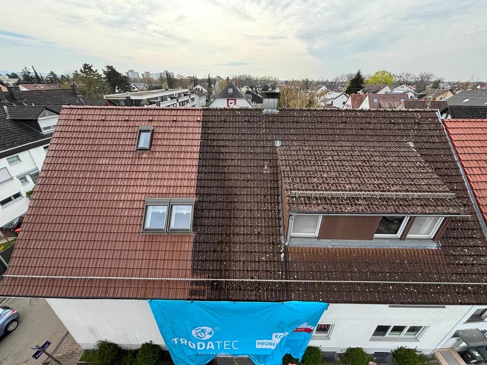 Drohnenaufnahme eines Mehrfamilienhausdachs während der Dachreinigung: links gereinigte rote Ziegel, rechts noch dunkel bewachsene Fläche